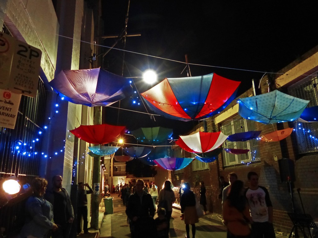 Schirmen hängen kopfüber an Wäscheleinen über der Straße. Darüber hängt eine blaue Lichterkette