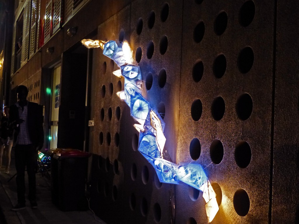 Ein aus weiße Fäden gesponnenes Kunstwerk das von innen beleuchtet wird, hängt an einer Wand